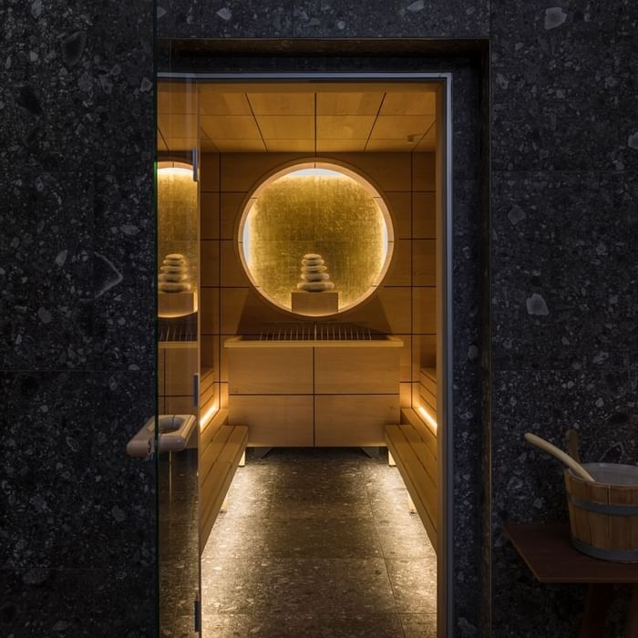 Sauna featuring wooden benches, circular window, and soft lighting on stone walls at Falkensteiner Hotels & Residences