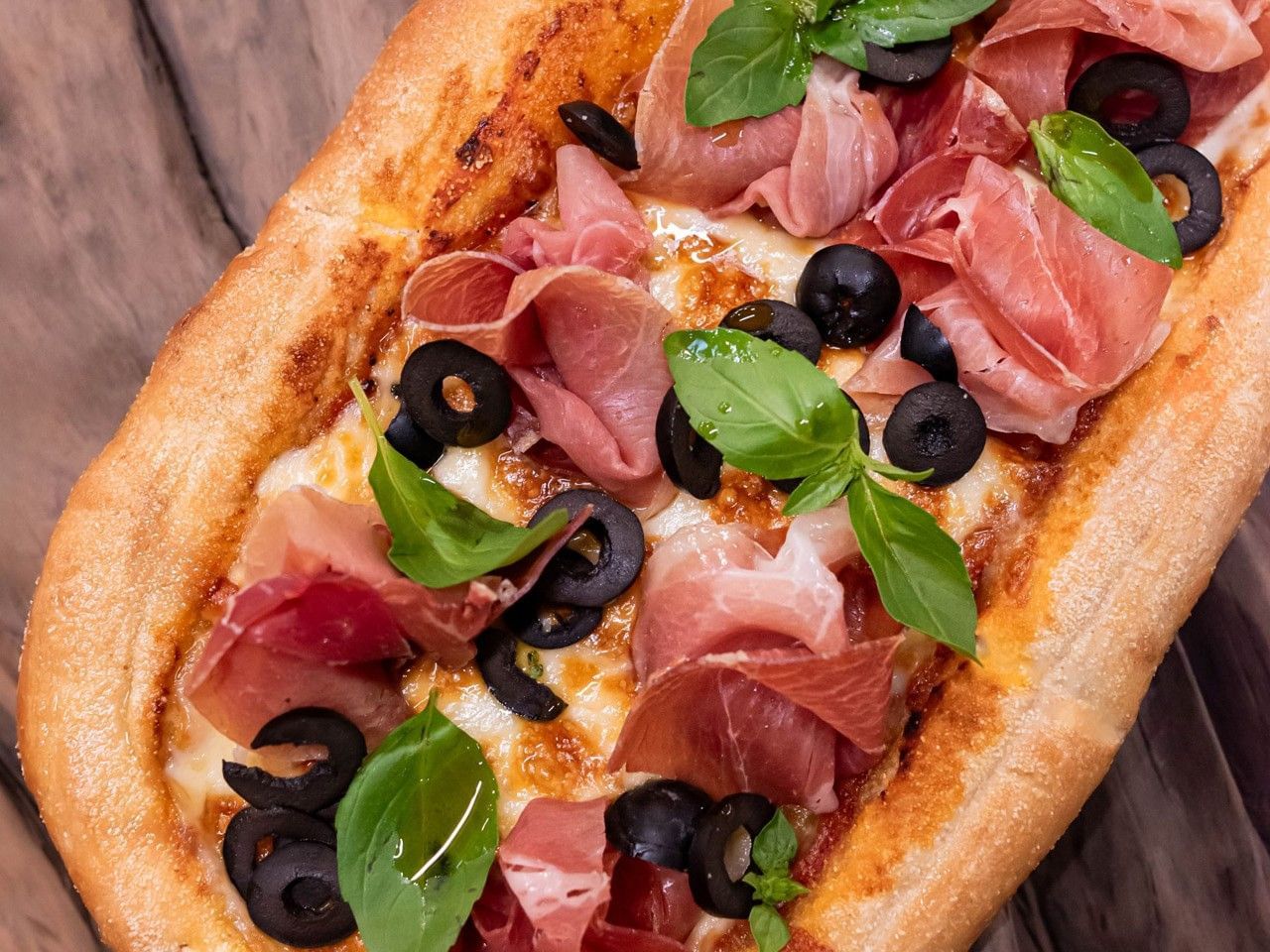 Pizza con jamón serrano y aceitunas, decorada con albahaca, servida en una tabla de madera en el hotel HCC Costanera Concepción.