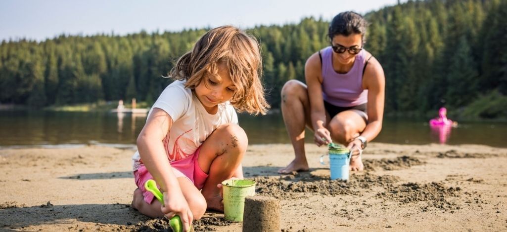 Child building a sandcastle at Lost Lake in Whistler while an adult plays nearby on the beach.