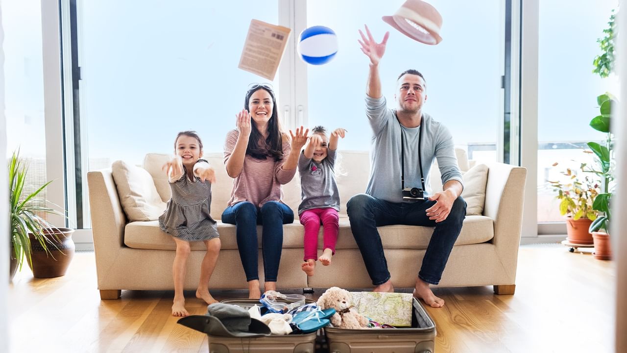 Family of four planning coast getaway with suitcase, throwing beach ball and hat in air.