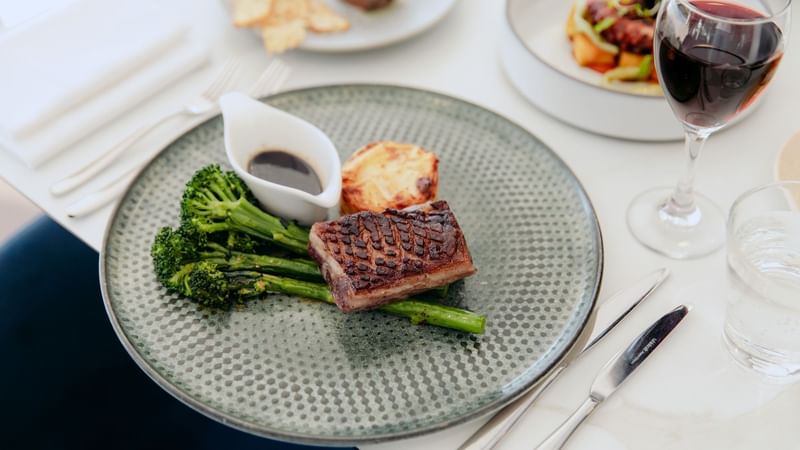 Plate with meat, broccoli, asparagus, sauce, knife, fork, and wine glass on a table with other dishes.