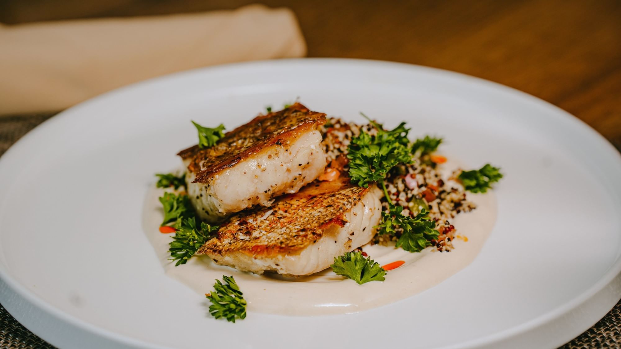 Plate with two pieces of fish, white sauce, and garnish of parsley and carrots on a wooden table.