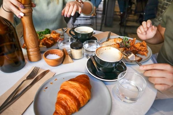 A close-up of two women at a table set with plates of breakfast food, a cappuccino, a croissant, and a wooden pepper grinder. 