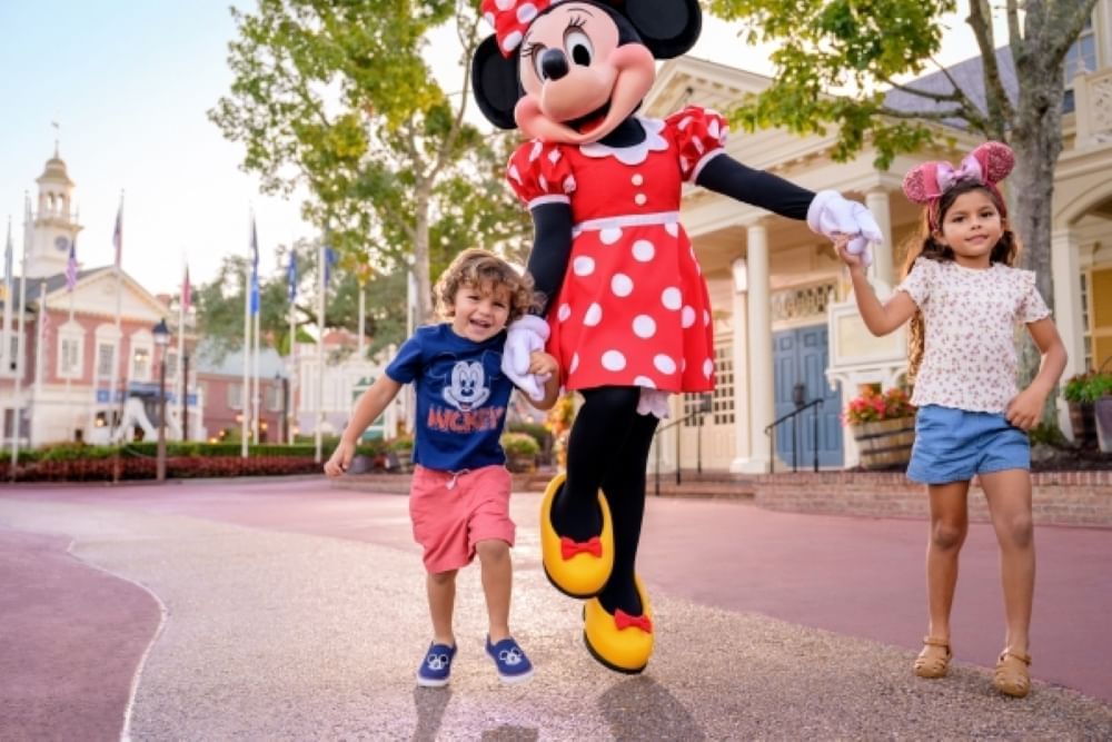 A young boy and girl hold hands with Minnie Mouse and walk through the streets of Magic Kingdom.