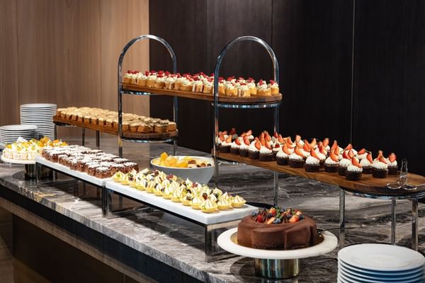 A dessert buffet with cakes, cupcakes, and fruit on a marble counter with stacks of plates.