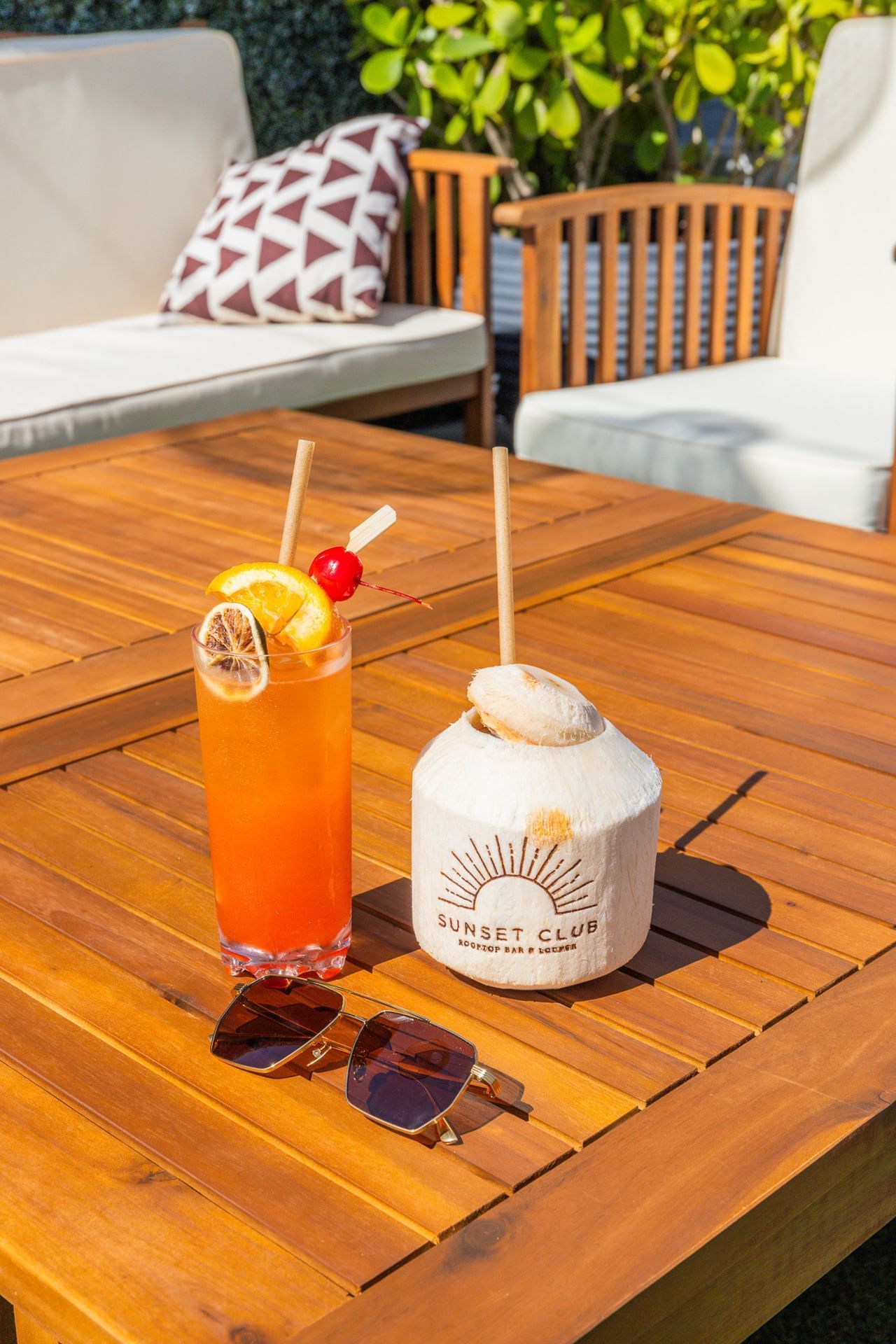 A wooden table with two drinks and sunglasses on it, with outdoor furniture in the background.