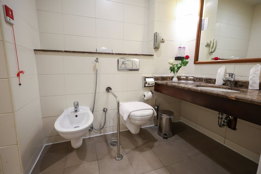Accessible room bathroom featuring a white bidet & toilet, with marble vanity and fresh red roses at Saja by Warwick Makkah
