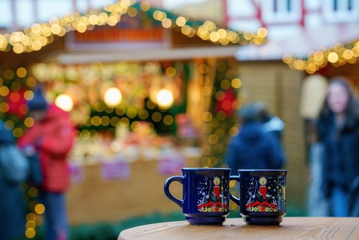 Two festive mugs sit on a table in front of a blurred Christmas market scene with people and lights.