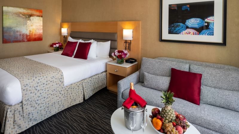 A hotel room with a bed, couch, fruit bowl, wine, and framed artwork on the walls.