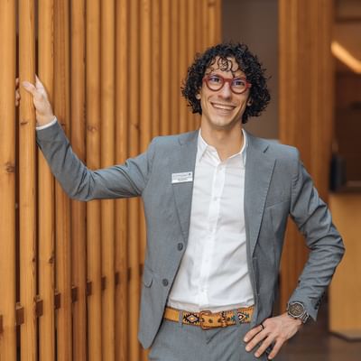 Lächelnder Mann mit Brille und grauem Anzug vor hölzernem Hintergrund.