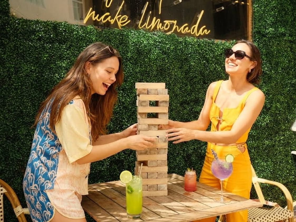 Two women playing a wooden block stacking game at an outdoor table with colorful cocktails during Labor Day at Limonada
