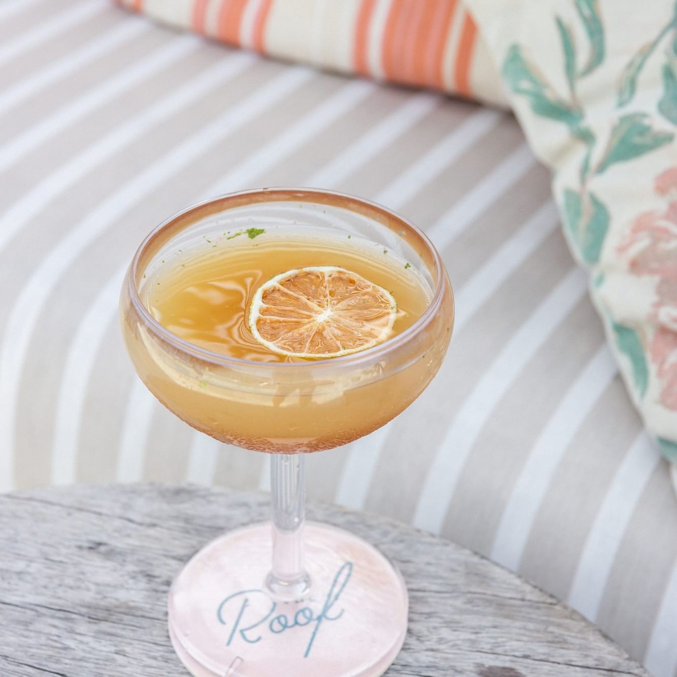 Golden cocktail garnished with dried citrus slice in coupe glass on weathered table with striped cushions for Martini Thursday