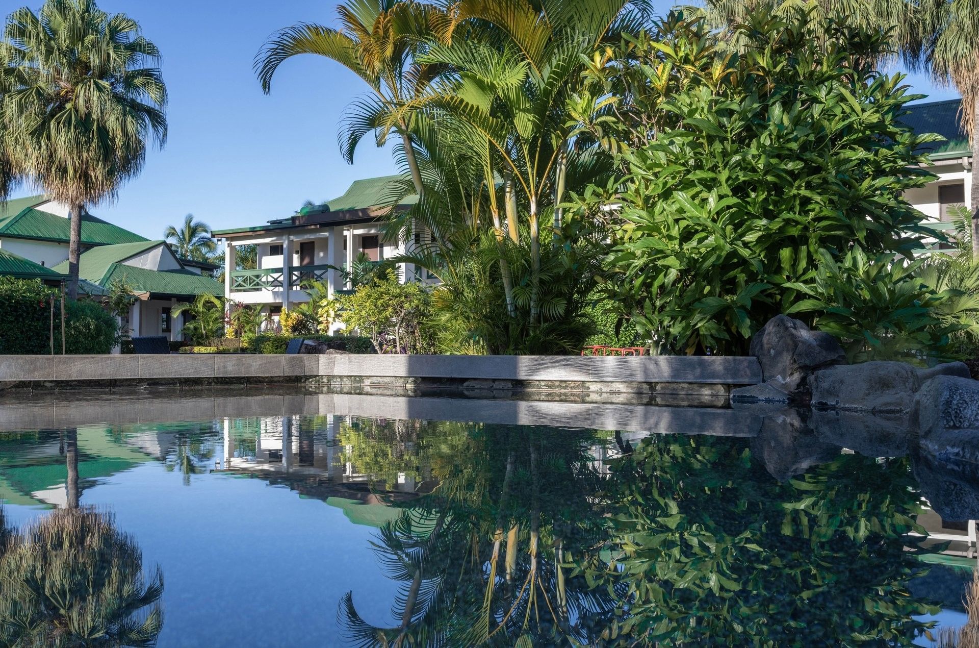 Lush palm trees reflected in a calm swimming pool by green-roofed buildings at TokaToka Resort Nadi Fiji