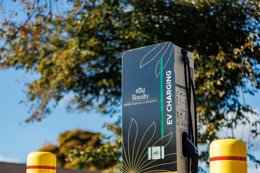 Black and gold EV charging station with a glowing green status light in the tree-lined parking lot at Rodd Royalty