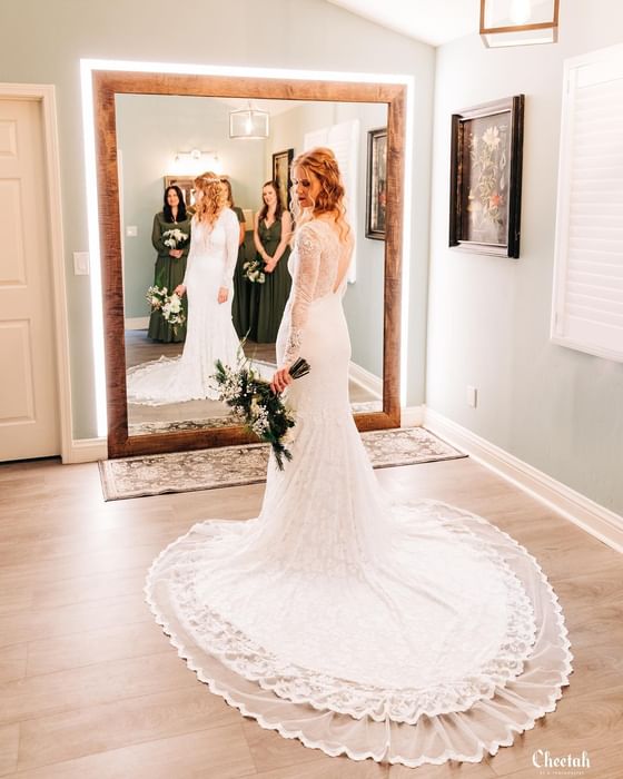 Bride in white lace gown with long train holding bouquet with bridesmaids in green dresses reflected in mirror at Cambria Pines Lodge