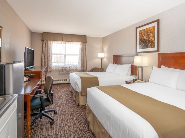 A hotel room with two beds, a work desk, a chair, a refrigerator, a sink, and a window.