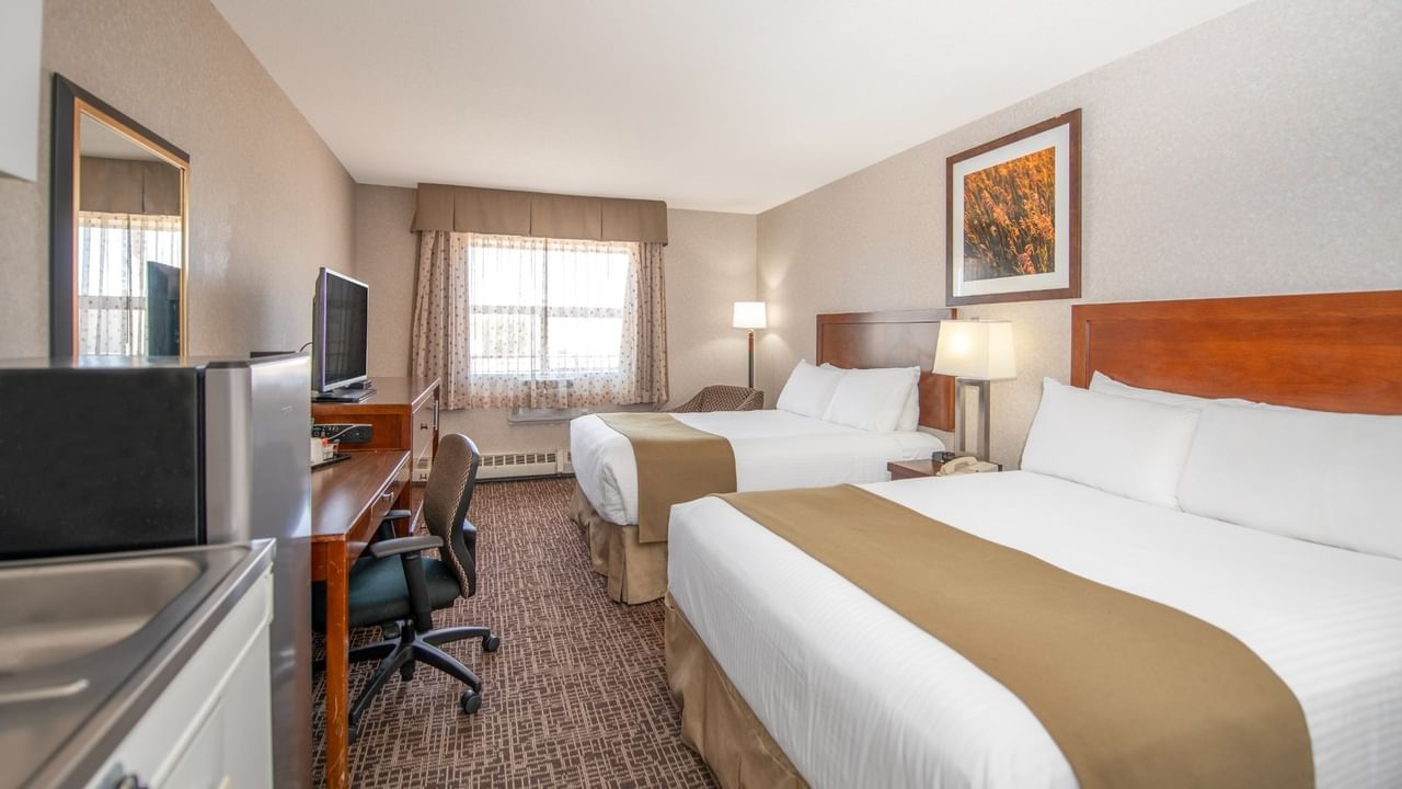 A hotel room with two beds, a work desk, a chair, a refrigerator, a sink, and a window.