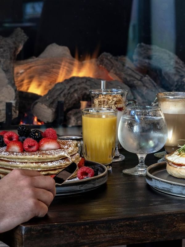 A breakfast spread with pancakes, orange juice, and a glass of water in front of a fireplace.