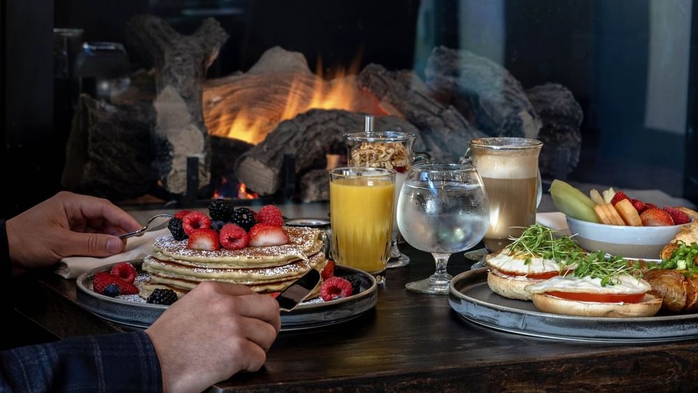 Stack of pancakes with berries by orange juice under the glow of a fireplace in In-Room Dining at Warwick Denver