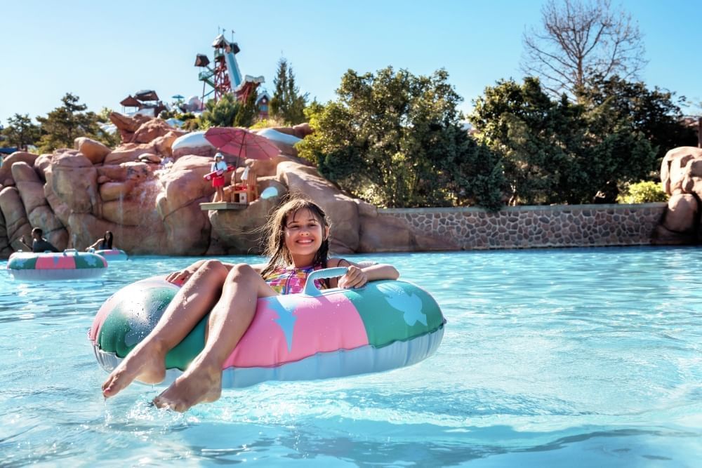 A girl sits and smiles in a colorful inner tube on the water with a large ski jump in the distance.