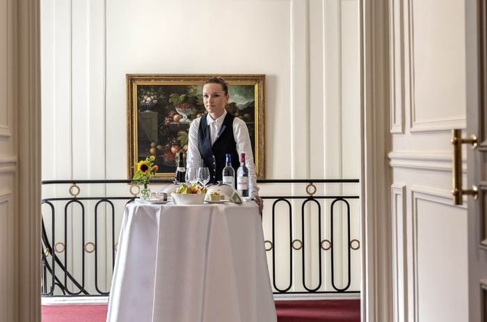 Membre du personnel en gilet noir derrière une table dressée avec tableau classique en arrière-plan à Hotel Westminster Warwick Paris