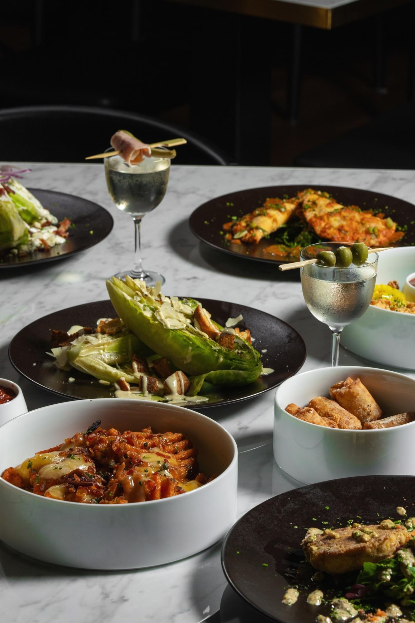 Marble table set with grilled romaine, pasta, and spring rolls, accompanied by cocktails and garnishes at Cypress & Oak