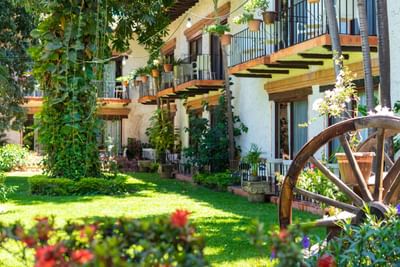 A building with balconies and potted plants, surrounded by greenery and a wagon wheel in the foreground.