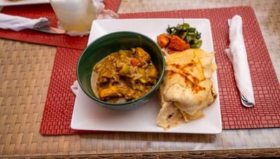 Green bowl of rich chicken curry served with a stack of folded roti and vegetables at Worthing Court Apartment