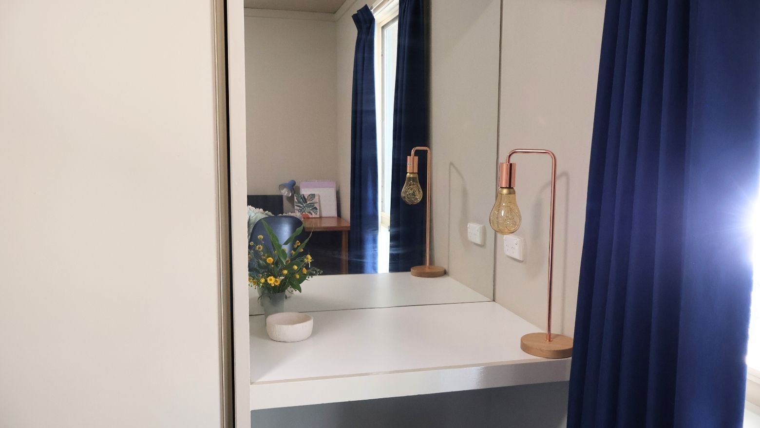 Mirror reflecting a room with blue curtains and a vase of flowers at La Trobe University Villas.