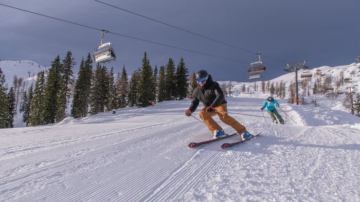 Two skiers on snowy slopes with ski lifts and trees in the background near Falkensteiner Hotel and Spa Carinzia