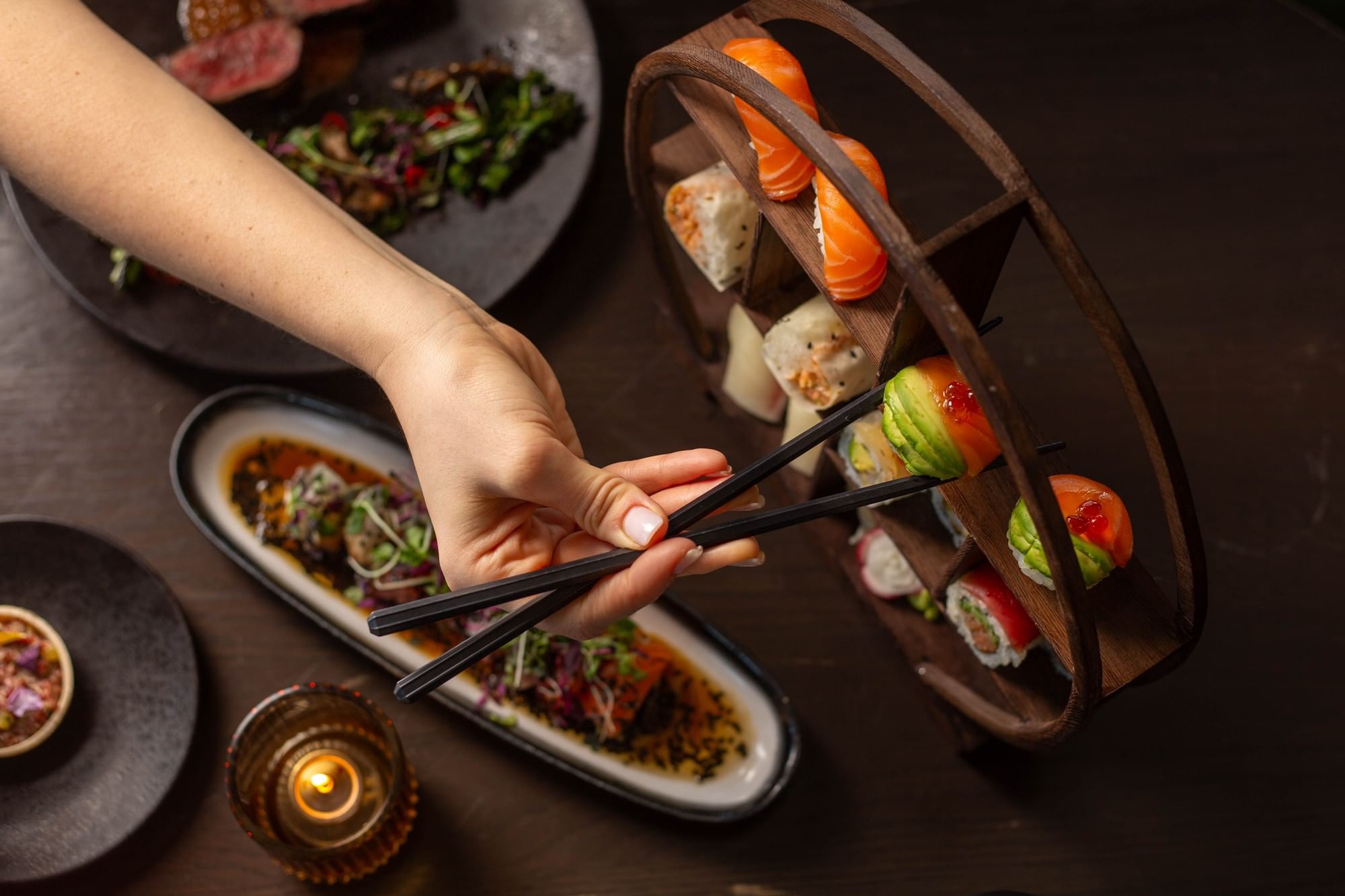 Hand holding chopsticks reaching Sushi and a plate of seared meat with garnishes in Valerie at Hotel X Toronto