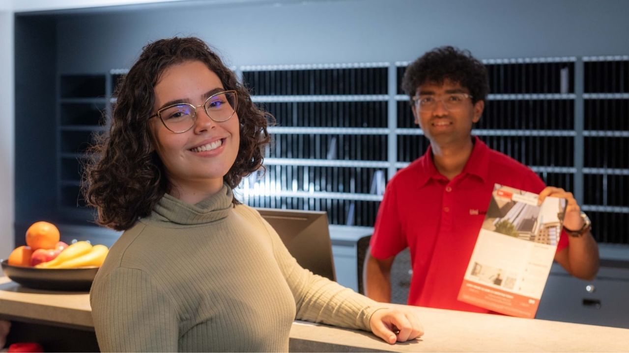A smiling resident with glasses stands behind a counter, next to staff holding a brochure.