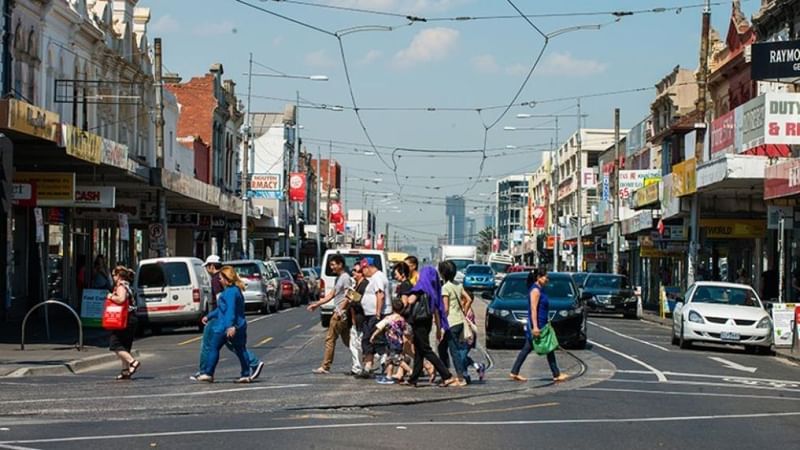 People crossing a street in Footscray (Melbourne) with shops on both sides and cars parked in front.