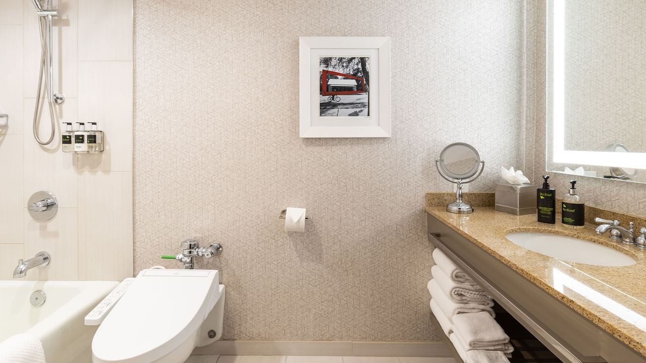 Hotel bathroom with toilet, sink, bathtub, framed picture, mirror, and folded towels on a shelf.