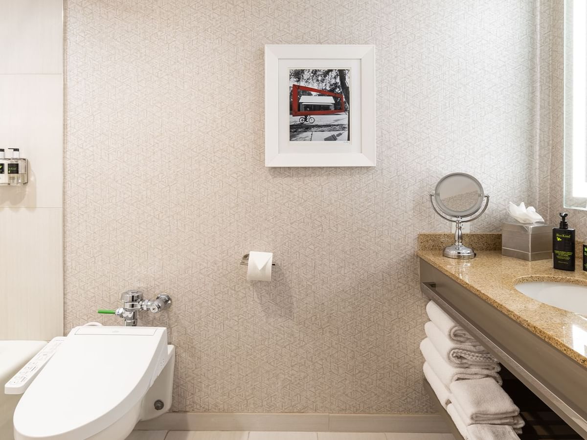 Hotel bathroom with toilet, sink, bathtub, framed picture, mirror, and folded towels on a shelf.