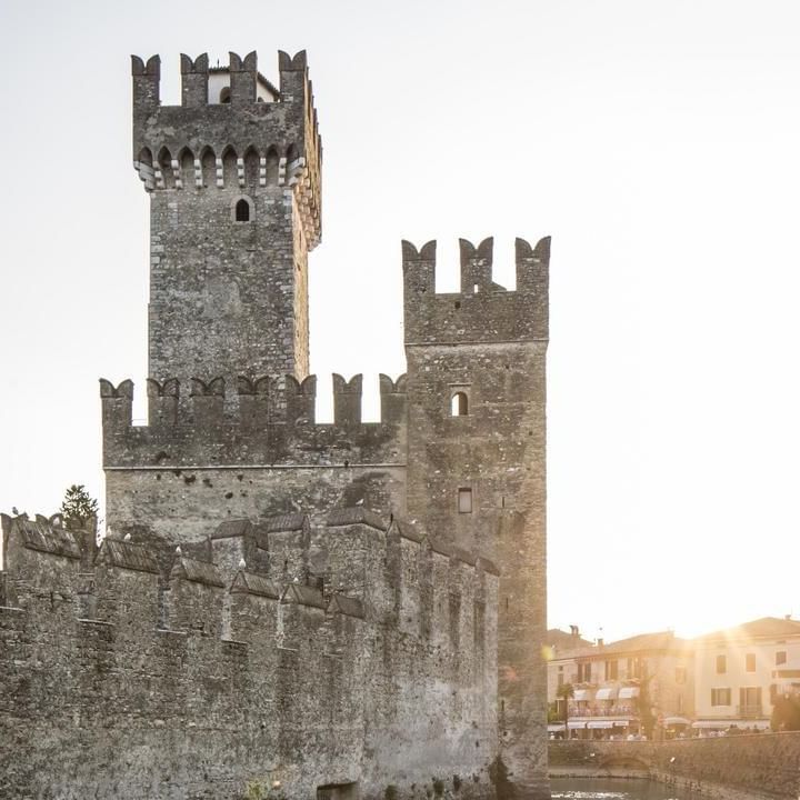Stone castle with two towers and walkway, surrounded by trees and buildings under a bright sky.
