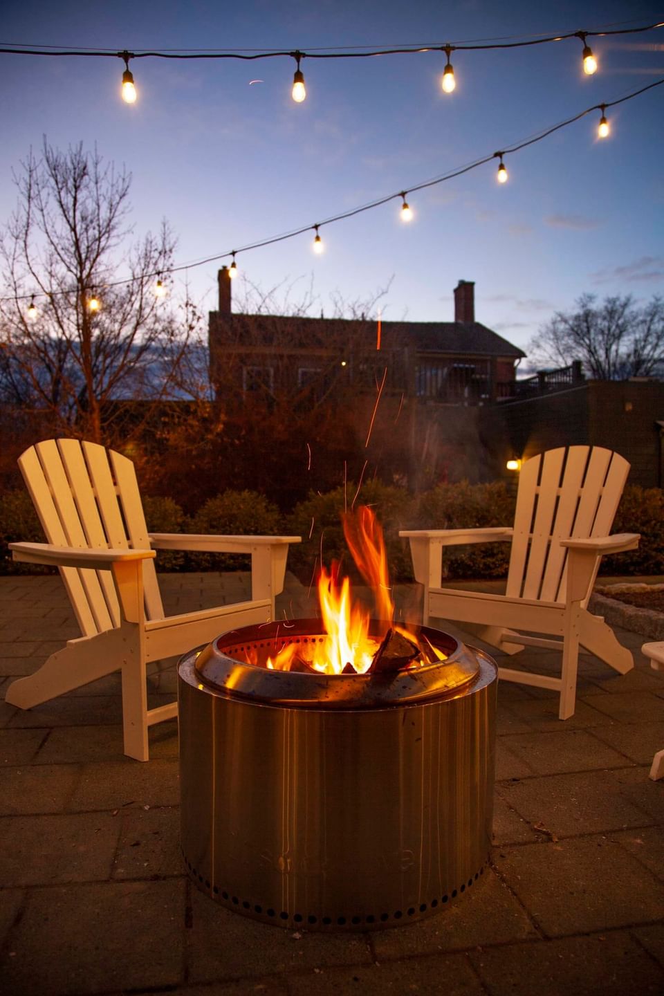 Warm glowing fire pit between white wooden Adirondack chairs under hanging string lights at night at Hotel X Toronto