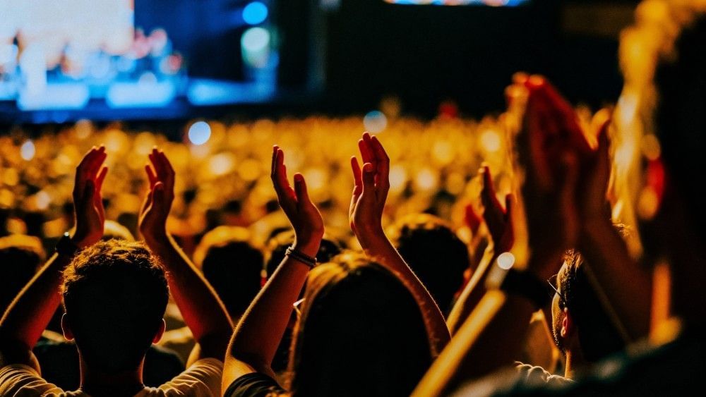 A silhouetted crowd of people at a concert with their hands raised above their heads as if clapping.