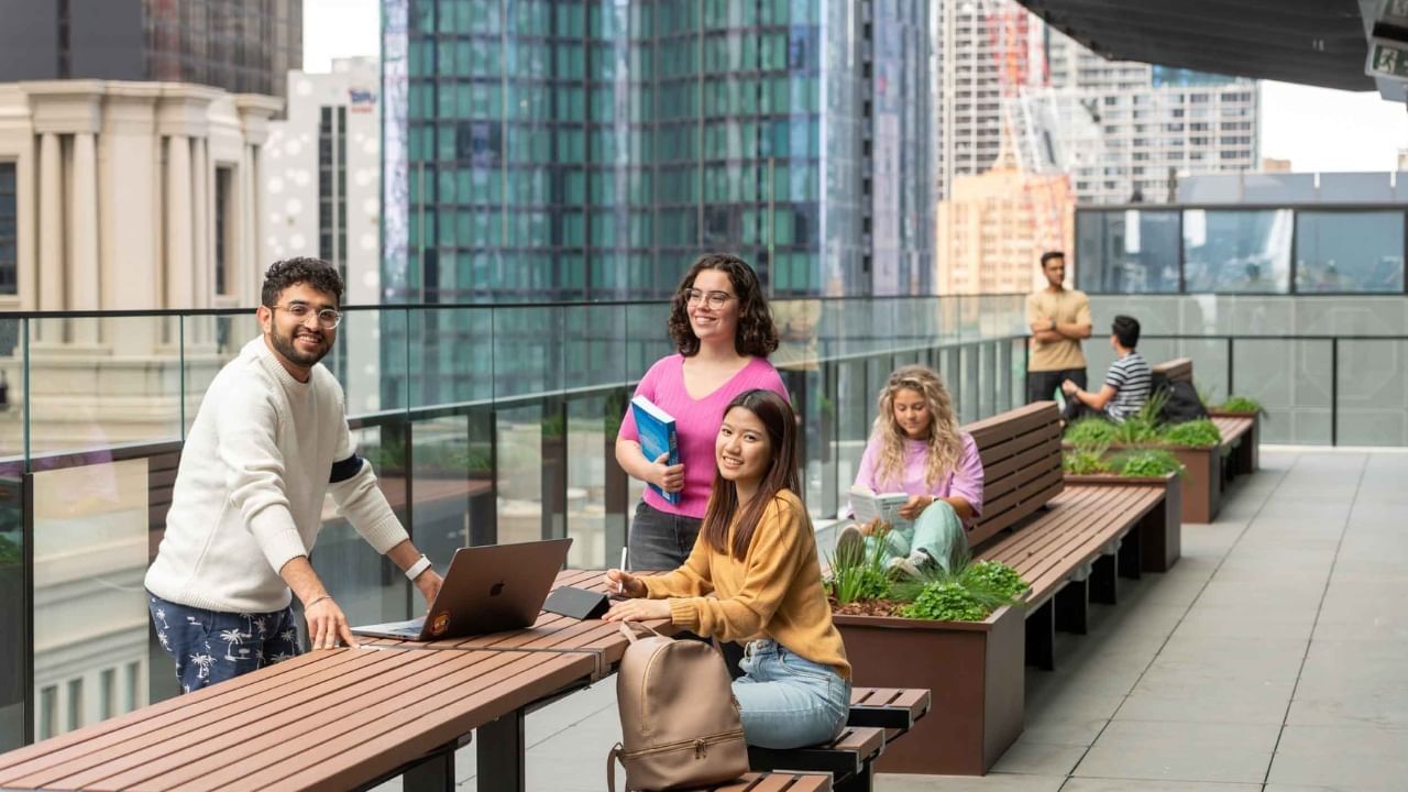 Group of students working and socializing on an outdoor rooftop with city skyline in background.
