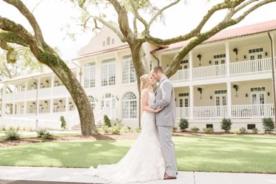 A bride and groom embracing on the lawn in front of the historic wedding venue and live oak trees at Centennial Plaza Resort