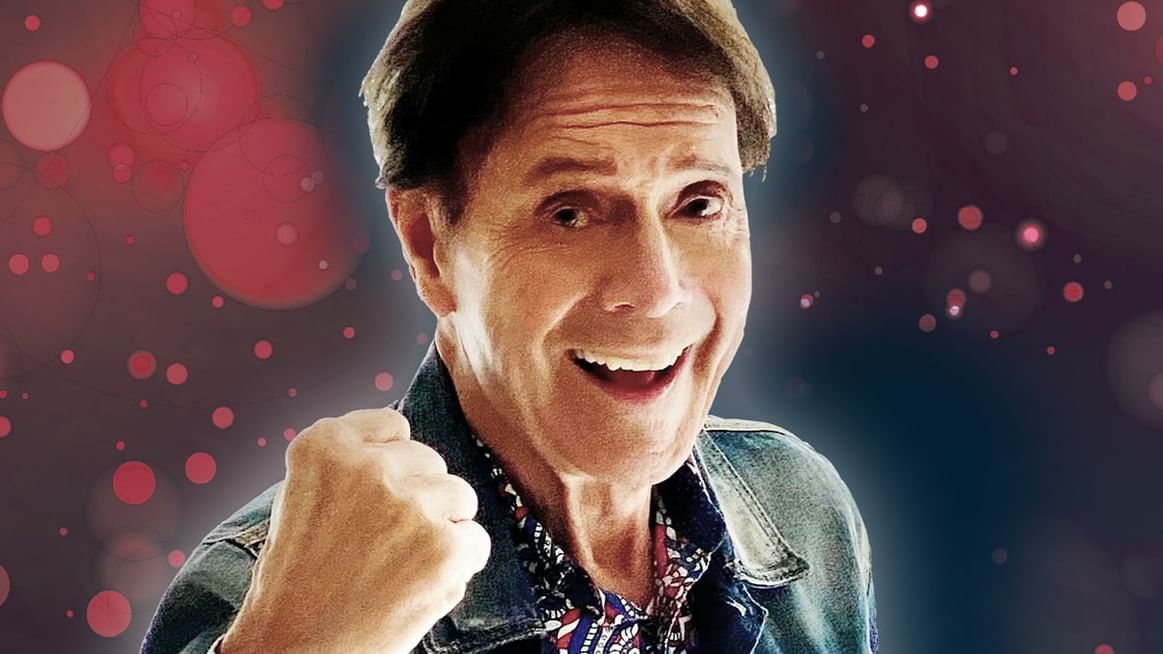 Sir Cliff Richard performing live on stage, smiling and holding a microphone, backed by vibrant stage lights