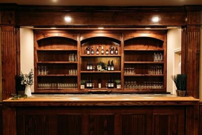 Wooden bar counter with bottles, glasses, and plants in a room with beige walls and dark brown accents.
