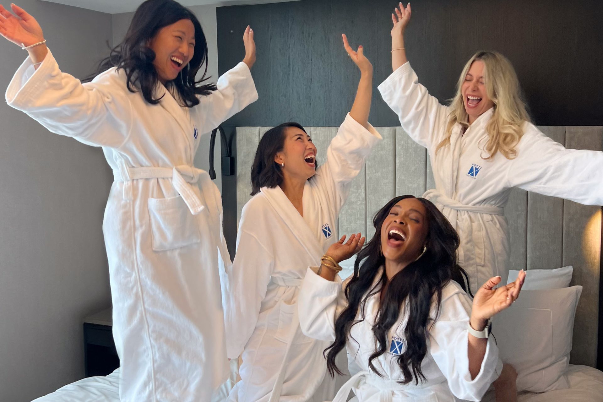 Four women in white robes laugh in a hotel room.