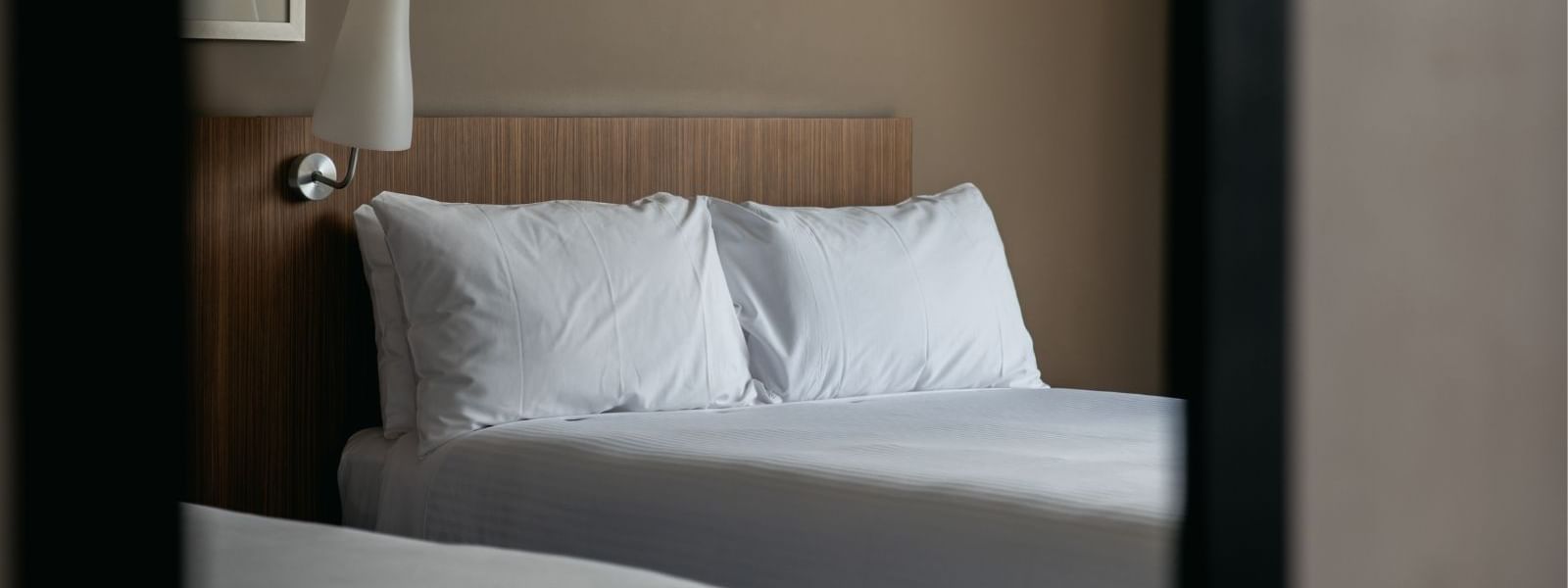 View of a hotel room with two beds, white bedding, and a wall-mounted lamp above the headboard.