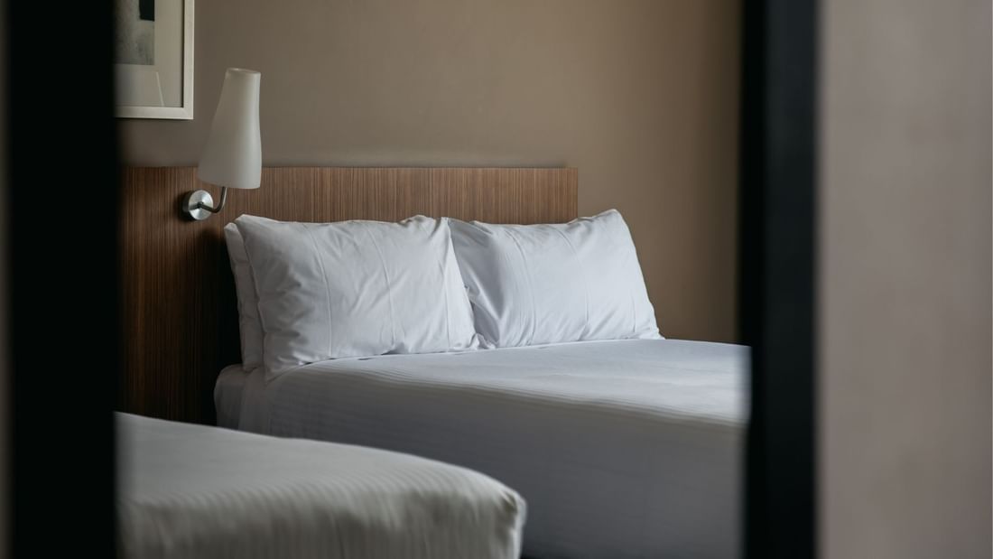 A hotel room with two beds, white linens, and a lamp mounted on the headboard.