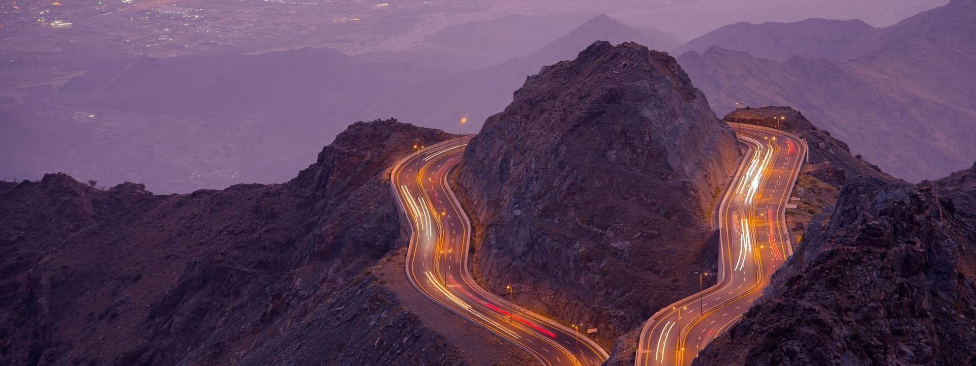 Hada mountain road illuminated by vehicle lights, framed by rugged terrain and a hazy valley near Warwick Hotels & Resorts