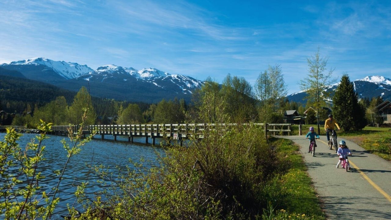 Family biking on Whistler Valley Trail with mountain view and wooden bridge.