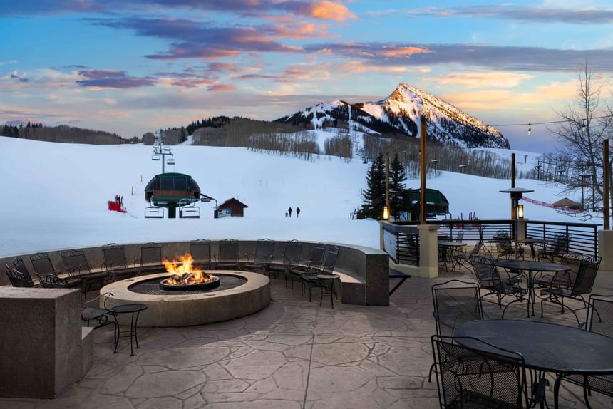 Outdoor seating area of the Elevation Hotel with fire pit, tables, and chairs, overlooking snowy mountain at sunset.