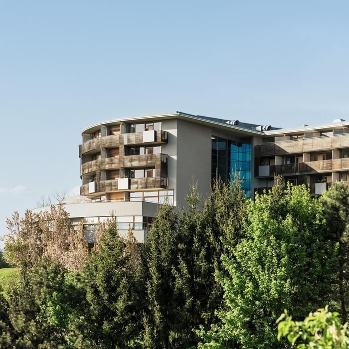 Modernes Gebäude mit Holzbalkonen und blauen Fenstern, umgeben von Bäumen und einem klaren Himmel.