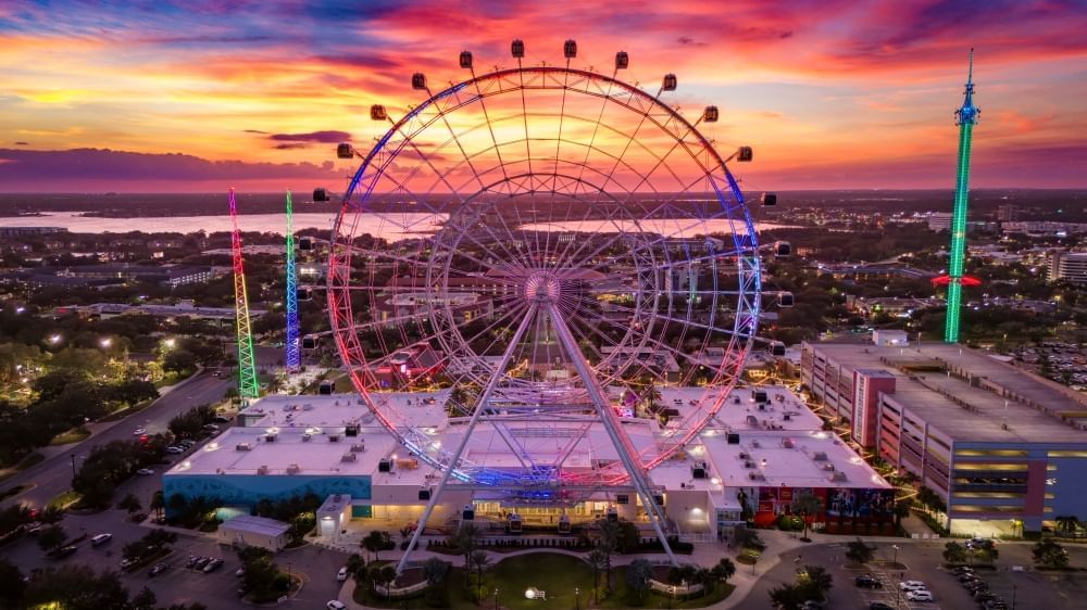 A large observation wheel surrounded by neon lights and attractions overlooking an orange and blue sunset sky.